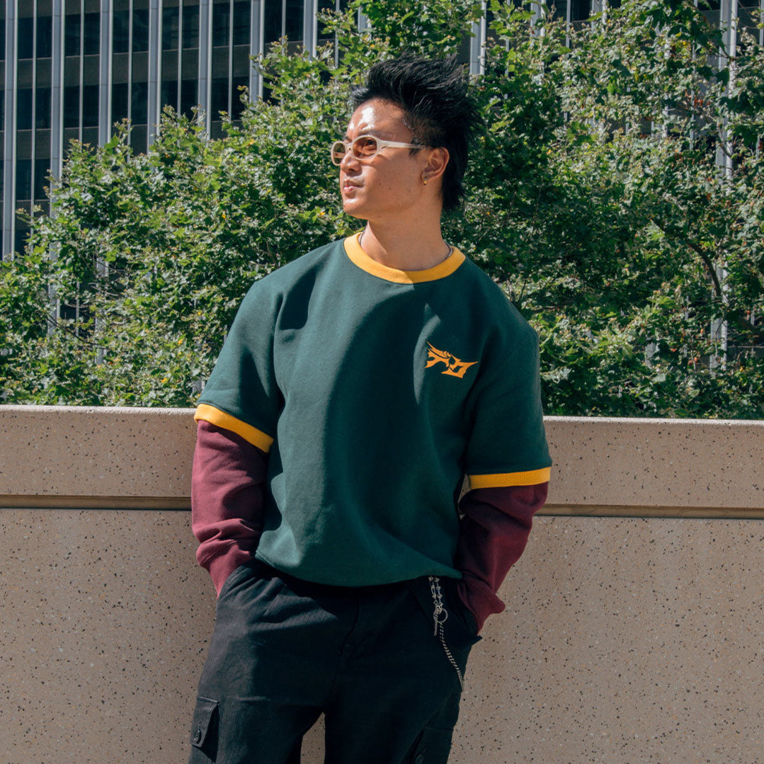 Man wearing a green and maroon sweater with a logo, standing outdoors with trees and buildings in the background.