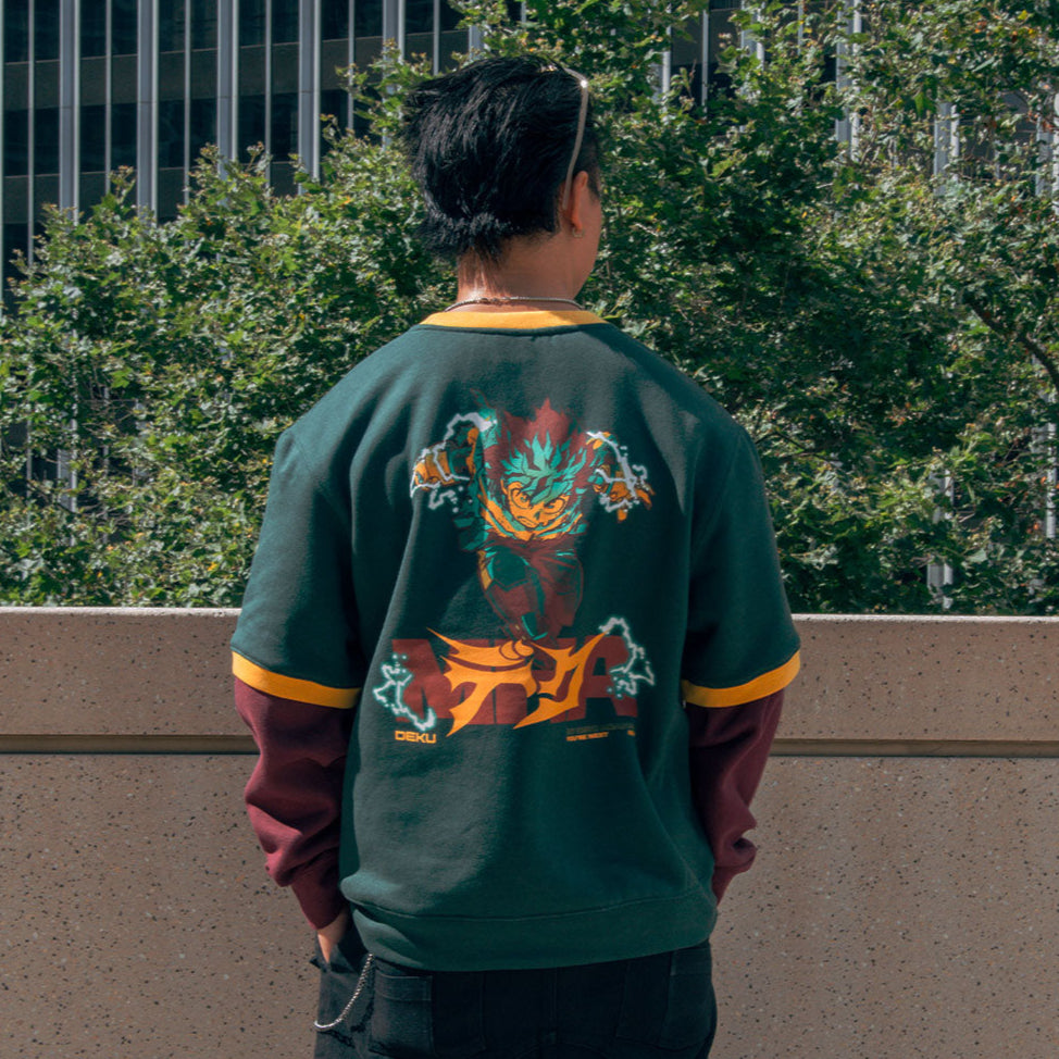 Person wearing a green varsity jacket with a colorful design on the back, standing in front of a building with trees.